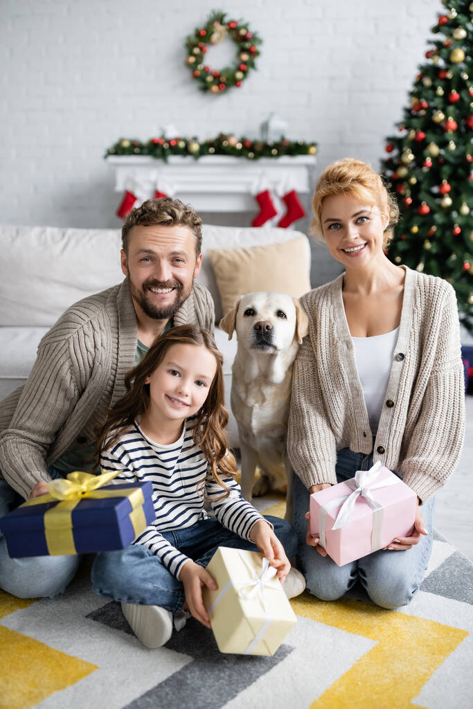 Positieve familie met geschenken en kijken naar camera in de buurt van labrador thuis  - Foto, afbeelding