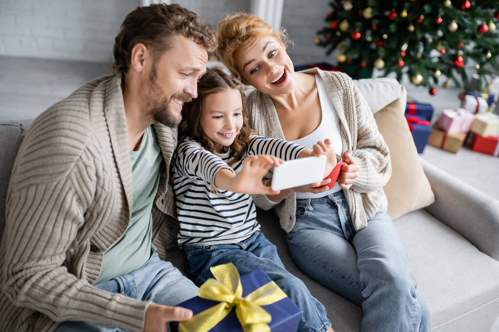 Chica sonriente tomando selfie en el teléfono inteligente cerca de los padres con la taza y el regalo de Navidad en casa  - Foto, imagen