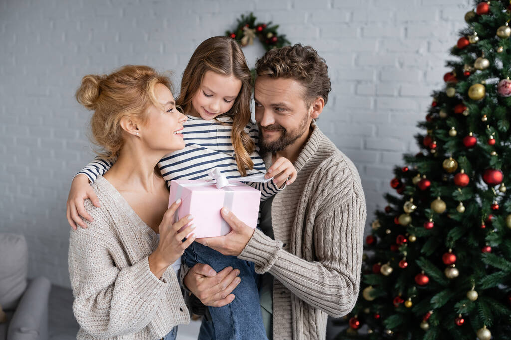 Sorrindo homem segurando presente e filha perto da esposa e árvore de Natal em casa  - Foto, Imagem