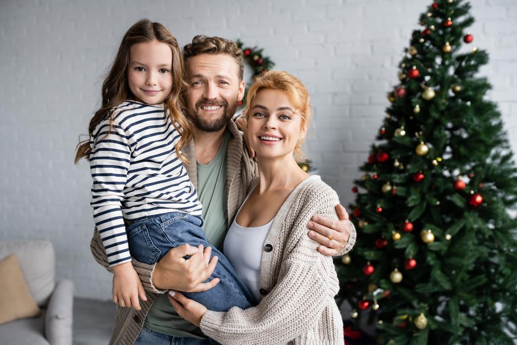 Un homme souriant étreignant sa femme et sa fille près d'un arbre de Noël flou à la maison  - Photo, image