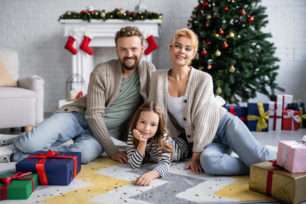 Uśmiechnięta rodzina patrząca w kamerę, siedząca obok konfetti i prezentów świątecznych na podłodze w domu  - Zdjęcie, obraz