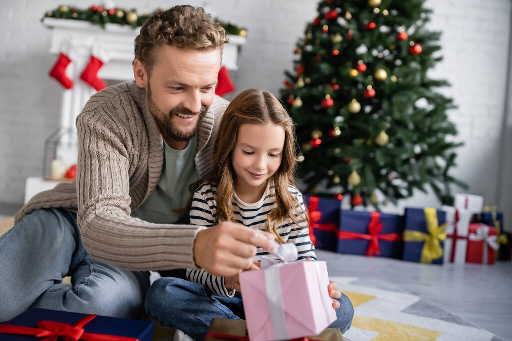 Homem positivo olhando para o presente perto da filha no chão durante o Natal em casa  - Foto, Imagem