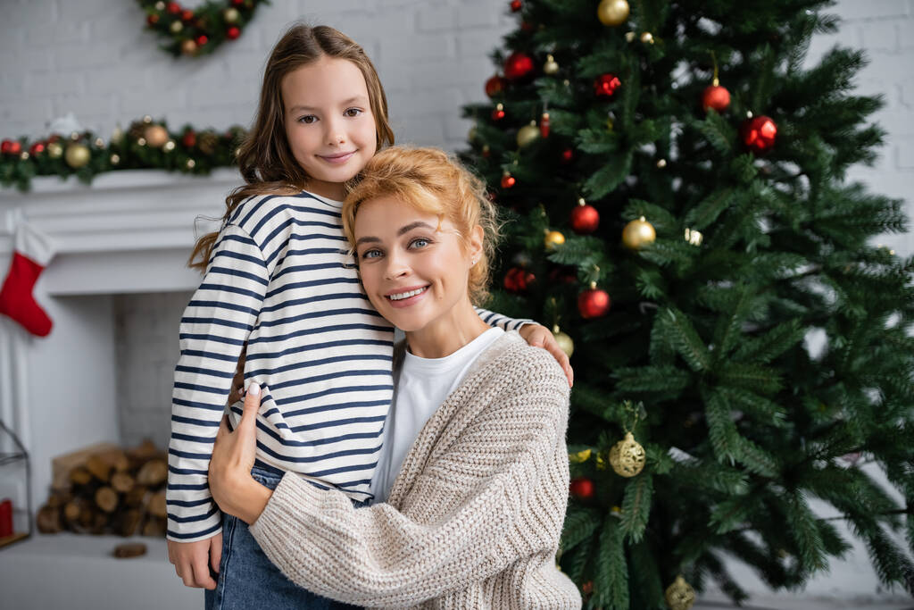 Sonriente mamá en punto cárdigan abrazando hija cerca de árbol de Navidad en casa  - Foto, Imagen