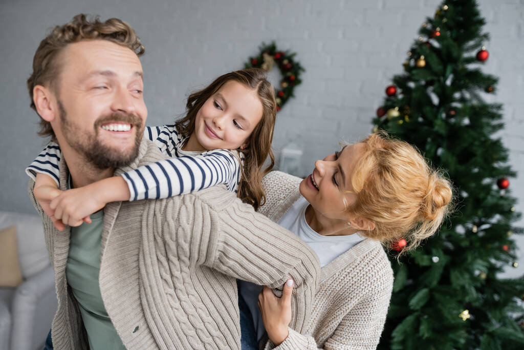 Χαρούμενο κορίτσι αγκαλιάζει τον μπαμπά κοντά στη μητέρα και θολή χριστουγεννιάτικο δέντρο στο σπίτι  - Φωτογραφία, εικόνα