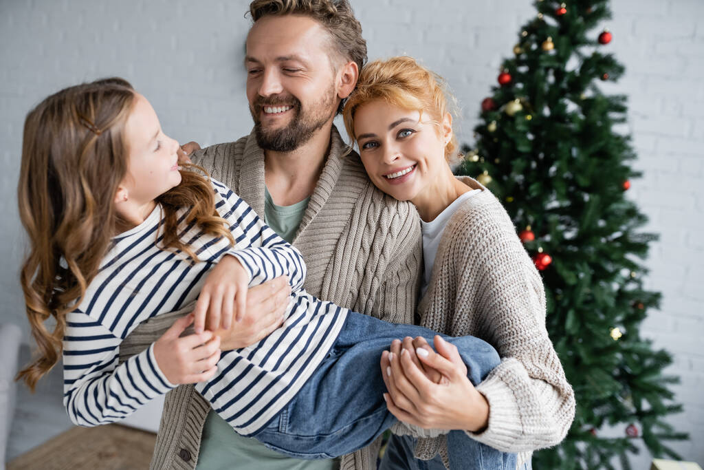 Lächelnde Eltern in warmen Strickjacken halten Tochter bei Neujahrsfeier zu Hause  - Foto, Bild