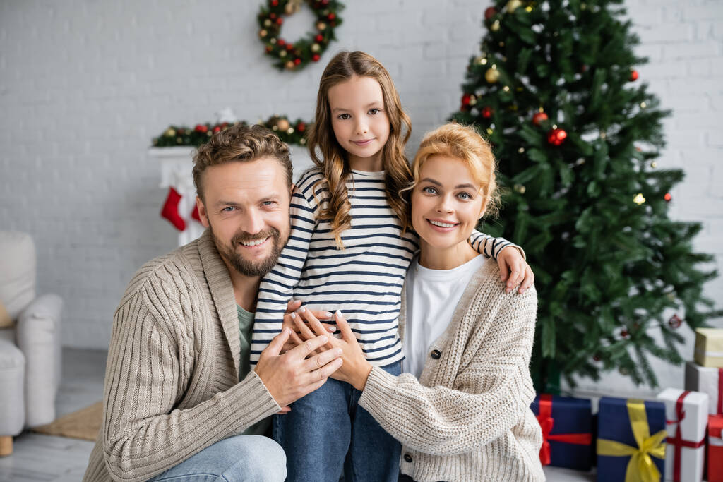 Веселая семья смотрит в камеру возле рождественской елки и размытые подарки дома  - Фото, изображение