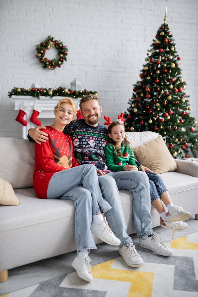 Familia positiva en suéteres abrazándose en el sofá cerca de la decoración borrosa de Navidad en casa  - Foto, imagen