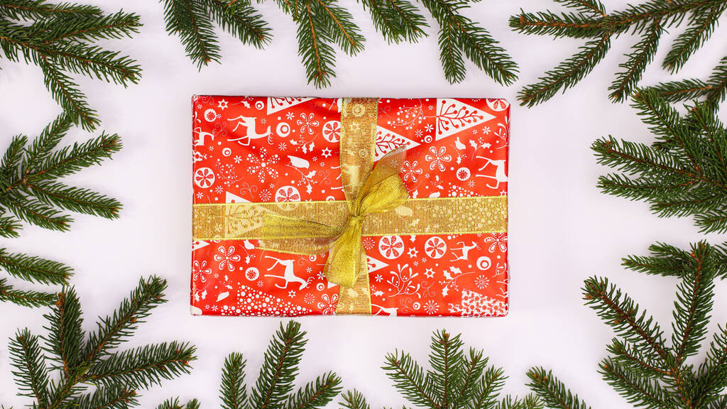Sosnowe gałęzie wokół czerwonego prezentu świątecznego ze złotą wstążką na białym tle. Układy płaskie - Zdjęcie, obraz