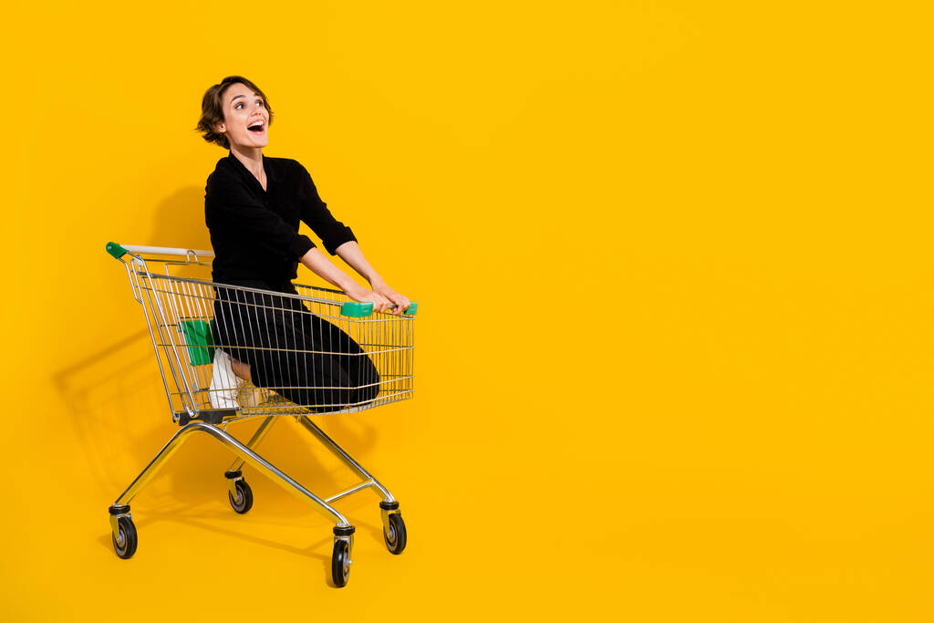 面白いの写真印象的な女性は空のスペース孤立黄色の背景を見てショッピングトレイに乗って黒のシャツを着用. - 写真・画像