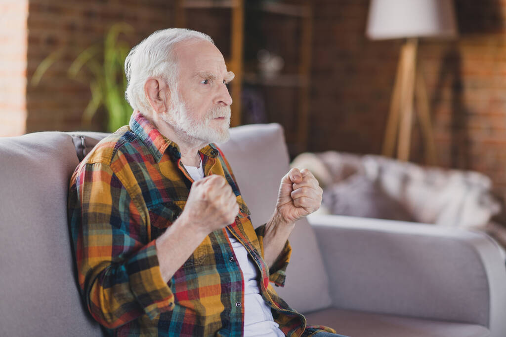 Photo of retired old man granddad grey hair raise fists watch bad news politics angry wear casual checkered look sitting indoors home room. - Photo, Image