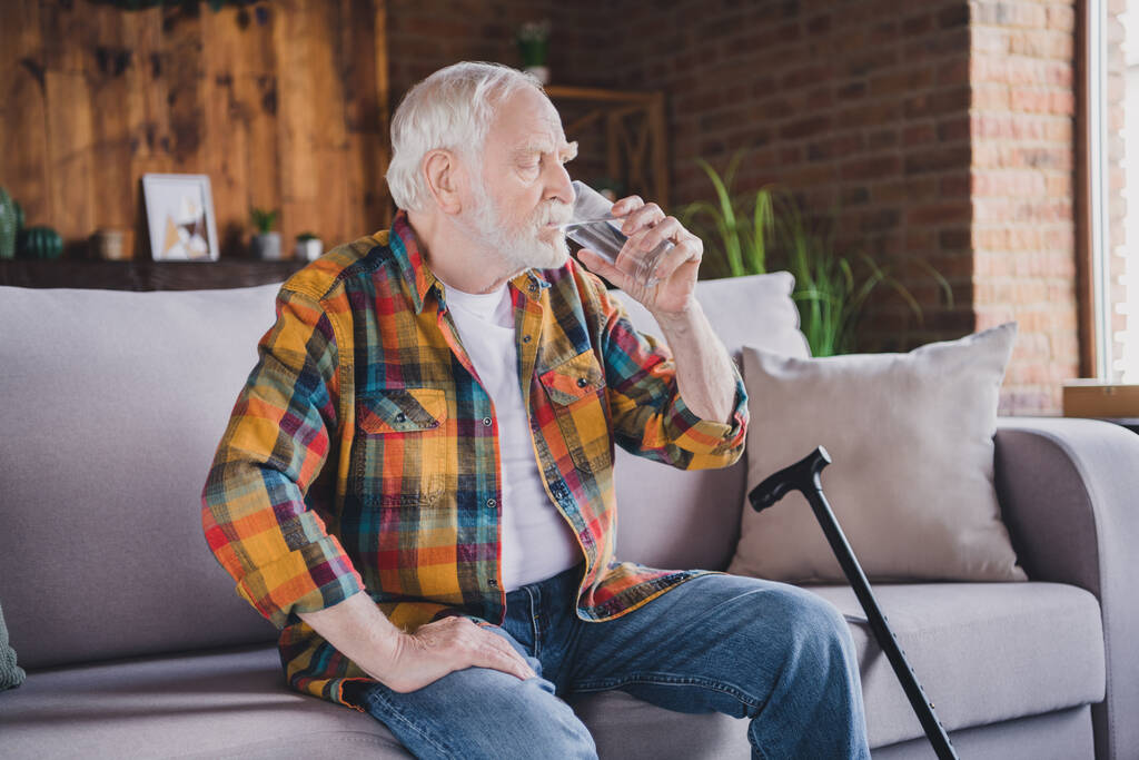 Φωτογραφία ενός σοβαρού όμορφου συνταξιούχου με καρό πουκάμισο που κάθεται στον καναπέ πίνοντας νερό στο εσωτερικό διαμέρισμα. - Φωτογραφία, εικόνα