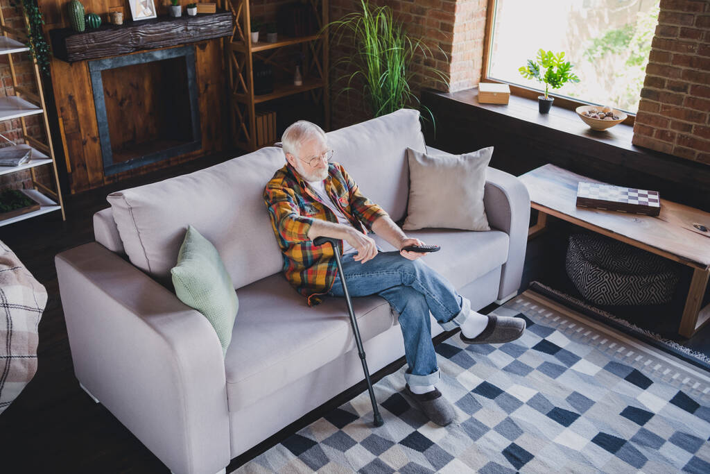 Фото гарного настрою чоловік пенсіонер одягнений у картату сорочку, яка дивиться фільм, що тримає прогулянкову тростину, сидячий диван у приміщенні будинку
. - Фото, зображення