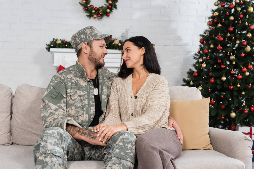 hombre en camuflaje abrazando esposa feliz mientras está sentado en el sofá cerca del árbol de Navidad decorado - Foto, imagen