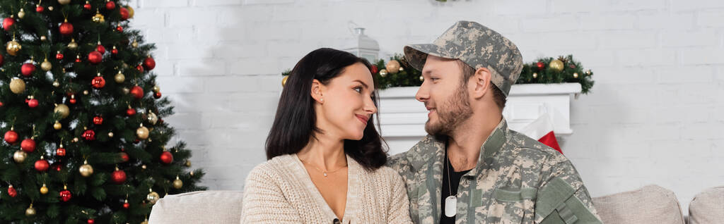 šťastná žena při pohledu na manžela ve vojenské uniformě v obývacím pokoji s vánoční stromeček, prapor - Fotografie, Obrázek