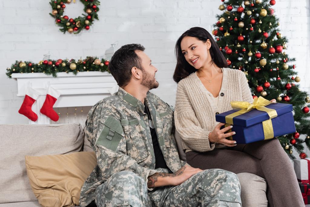 mujer alegre con caja de regalo mirando al marido en camuflaje cerca del árbol de Navidad decorado - Foto, Imagen