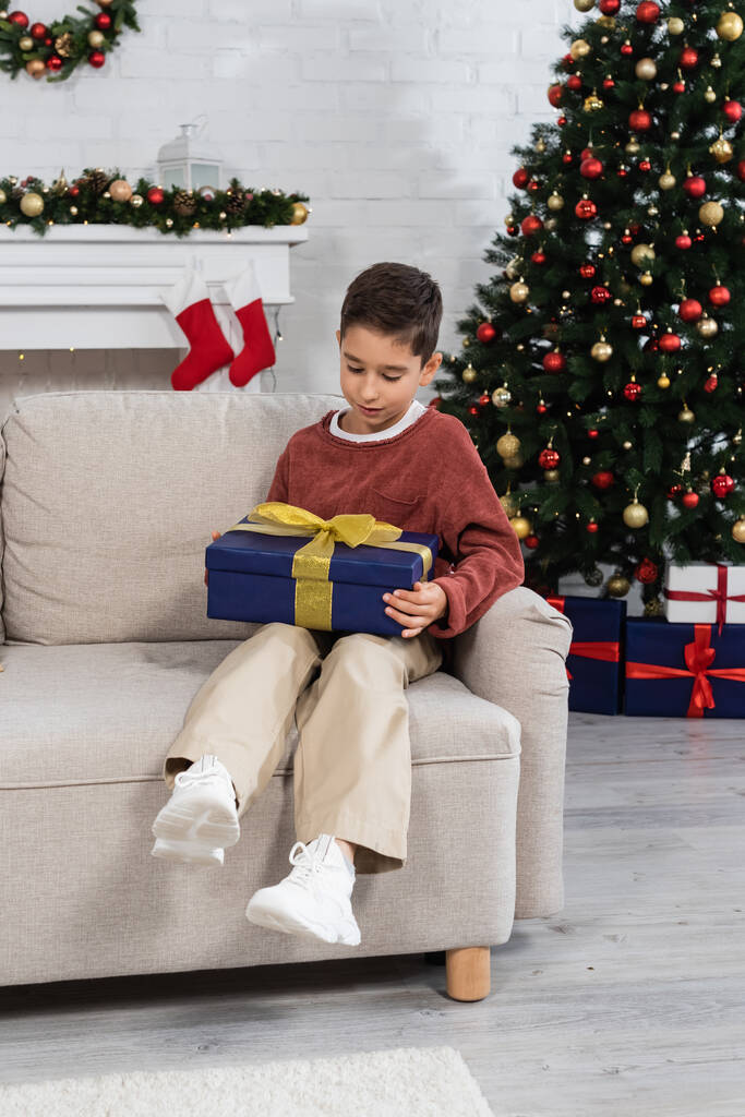comprimento total da criança com presente sentado no sofá perto da árvore de natal decorada em casa - Foto, Imagem