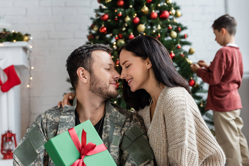 szakállas katona férfi és nő csukott szemmel mosolyog közelében ajándék doboz, míg a fia díszítő karácsonyfa  - Fotó, kép