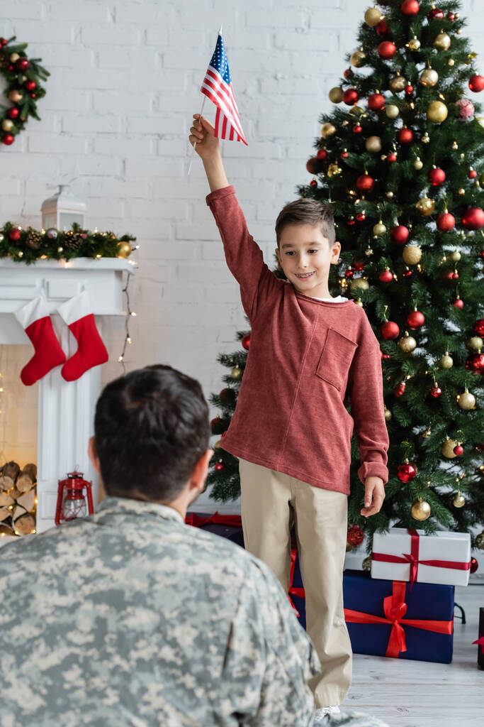 sorrindo menino patriótico de pé com bandeira dos EUA perto de árvore de natal e pai em camuflagem em primeiro plano borrado - Foto, Imagem