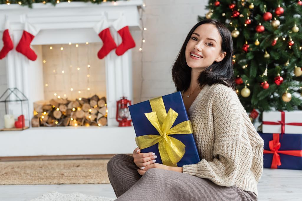 vidám nő ül ajándékdobozzal közel karácsonyfa és díszített kandalló otthon - Fotó, kép