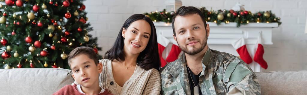 glücklicher Militärmann mit Frau und Sohn, der zu Hause in der Nähe des Weihnachtsbaums in die Kamera schaut, Banner - Foto, Bild