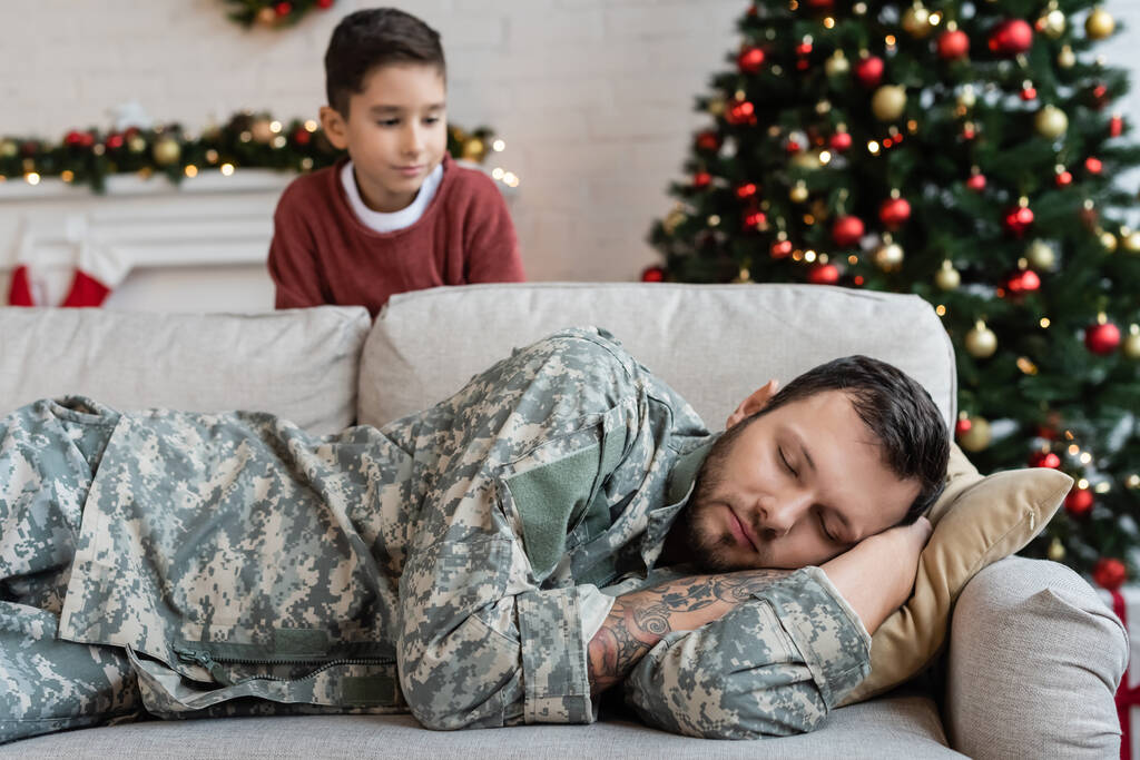 ぼやけた少年は疲れきったお父さんを見てるクリスマスの装飾付きリビングルームでソファで寝てる - 写真・画像