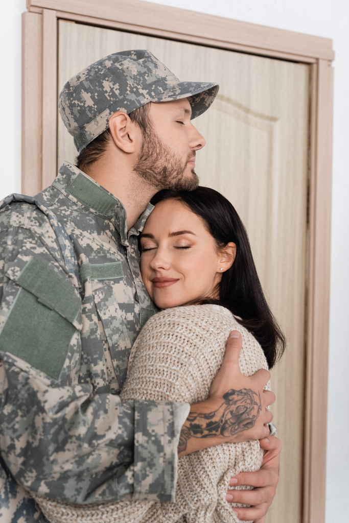 brodaty mężczyzna w mundurze wojskowym przytula szczęśliwą żonę podczas powrotu do domu - Zdjęcie, obraz