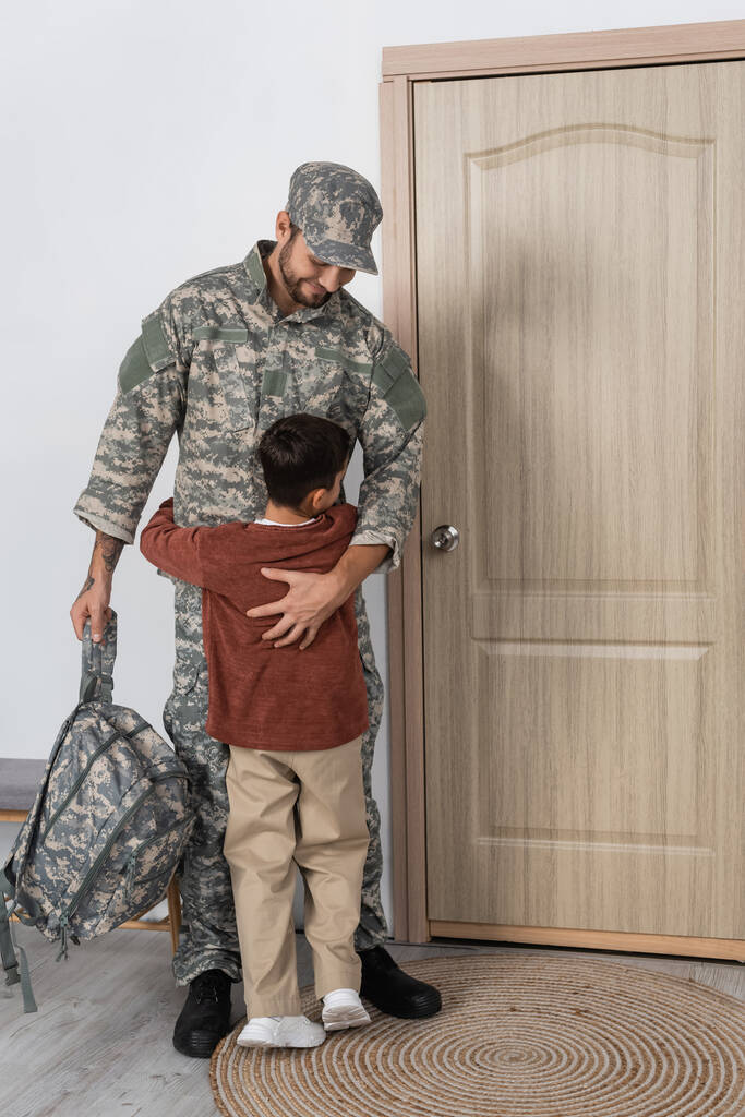 хлопчик обіймає тата у військовій формі, стоячи з рюкзаком біля вхідних дверей вдома
 - Фото, зображення