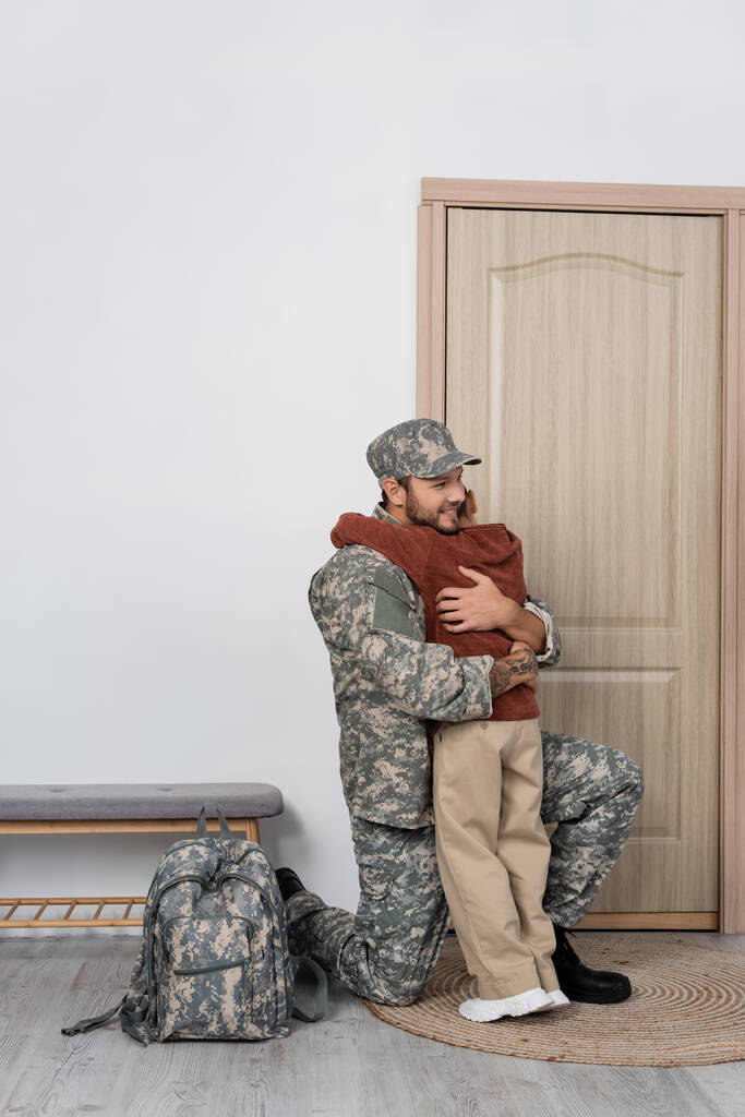 homem militar feliz abraçando filho perto de mochila e porta de entrada em casa - Foto, Imagem