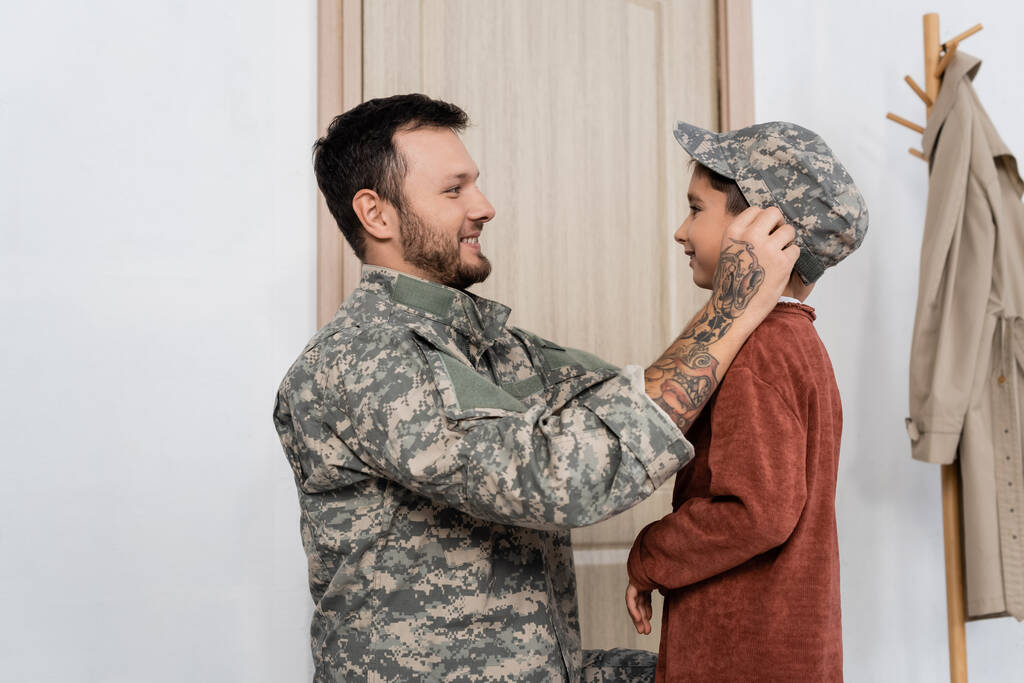 Seitenansicht eines bärtigen Mannes mit Militärmütze auf fröhlichem Sohn, der ihn zu Hause trifft - Foto, Bild