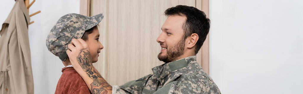 vista lateral de papá sonriente poniéndose gorra militar en el hijo cuando regrese a casa, pancarta - Foto, Imagen