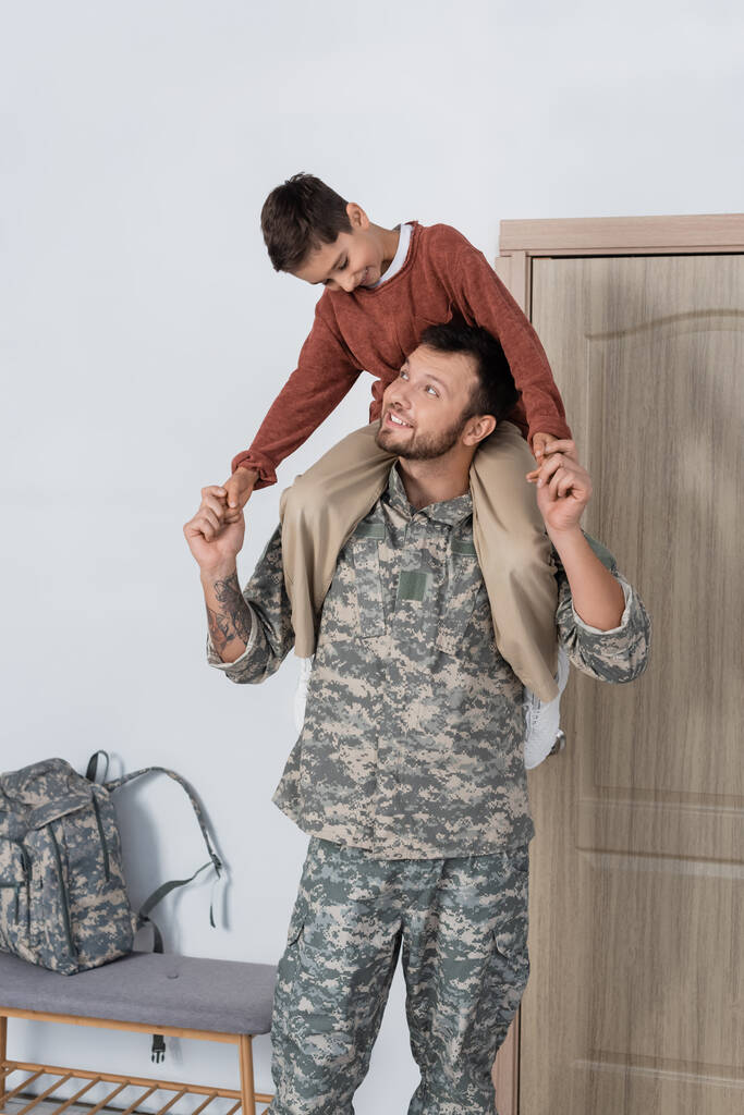felice ragazzo piggybacking padre in uniforme militare vicino porta d'ingresso a casa - Foto, immagini