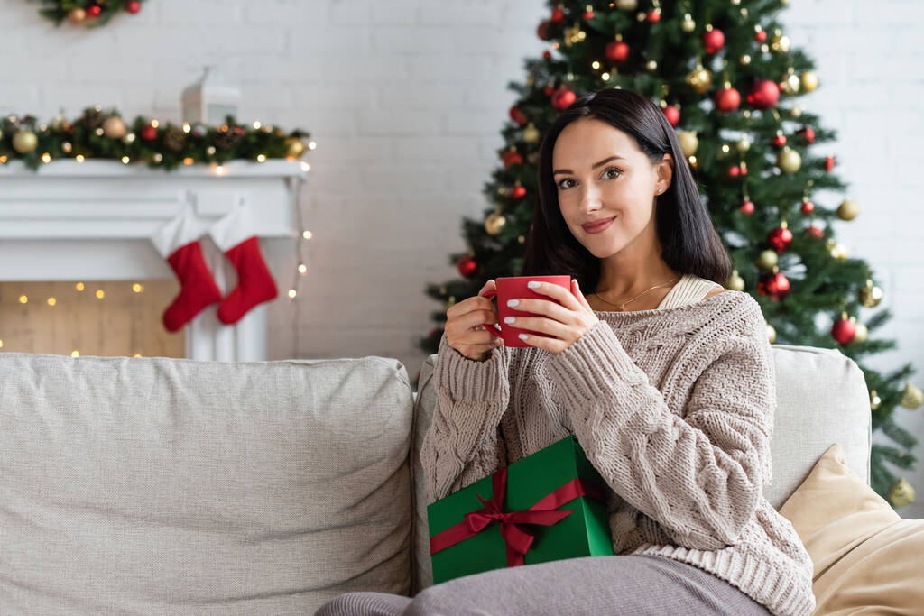 happy woman with cup of warm cocoa and gift box sitting on couch in living room with christmas decor - Photo, Image
