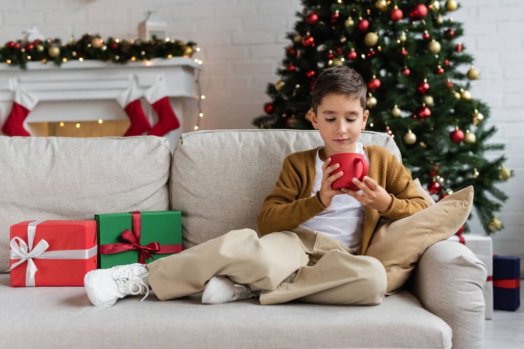 full length of kid holding cup of warm cocoa on couch near gift boxes and blurred christmas tree - Photo, Image