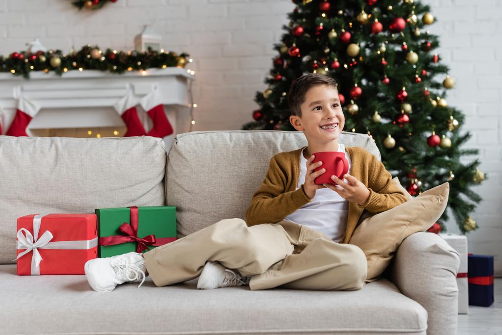 vrolijke jongen zitten op de bank met een kopje cacao en weg te kijken in de buurt van geschenkdozen en wazig kerstboom - Foto, afbeelding