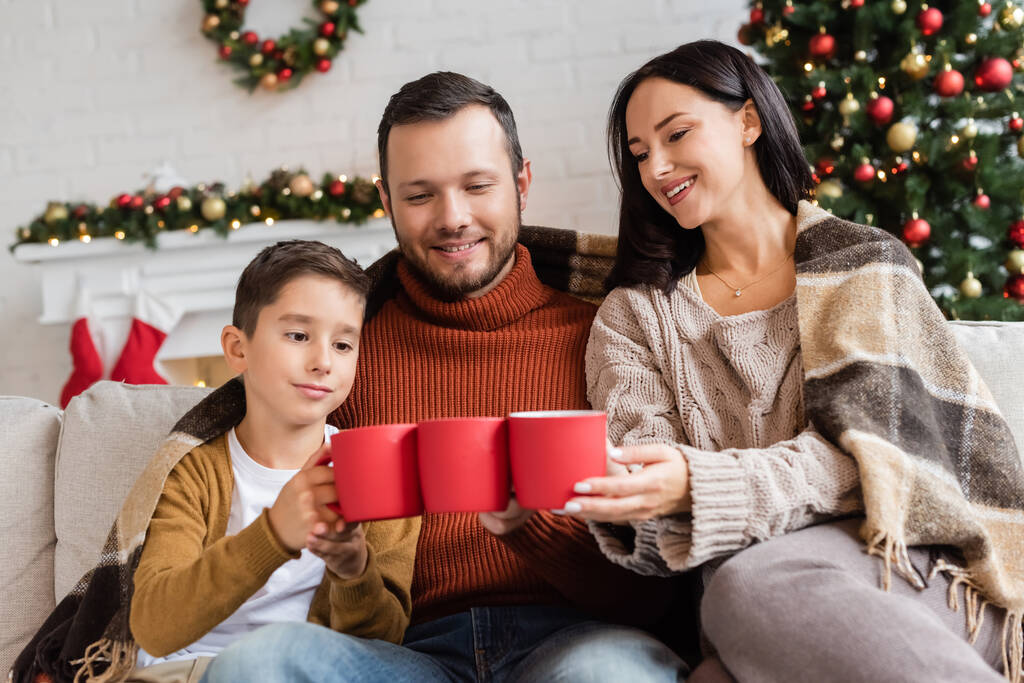 fröhliche Familie klappert Tassen mit Kakao auf Couch unter warmer Decke neben verschwommenem Weihnachtsbaum - Foto, Bild