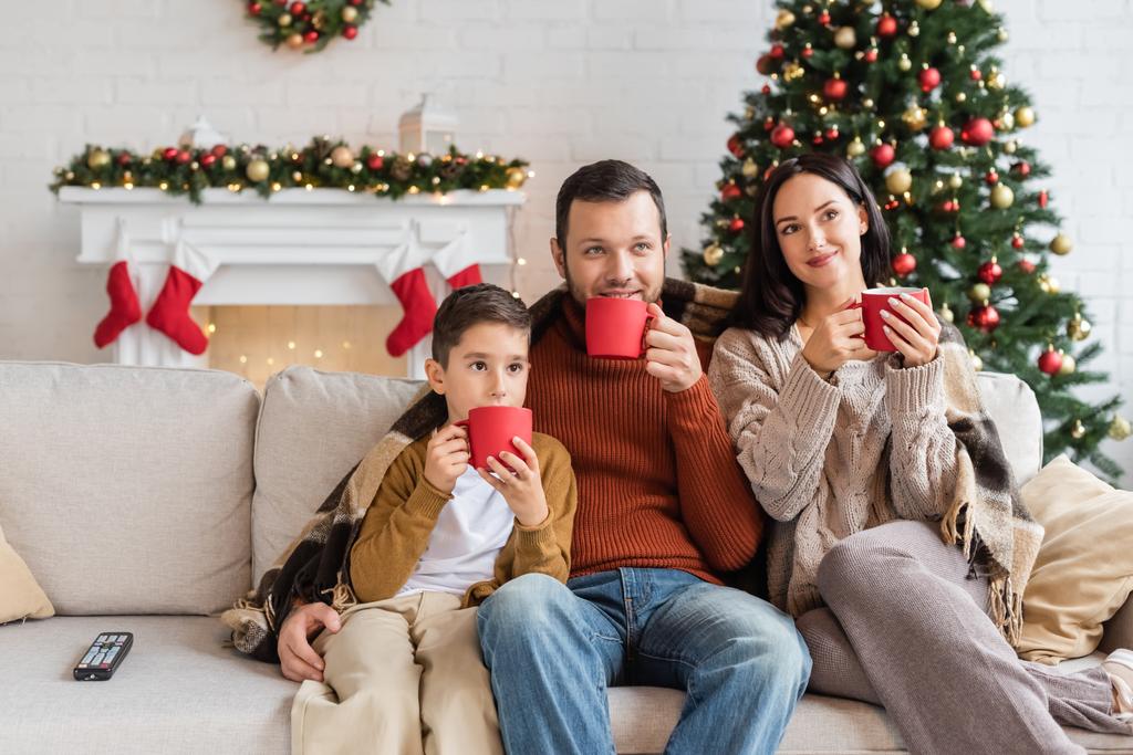 familia feliz beber cacao en el sofá debajo de la manta cerca del árbol de navidad borrosa - Foto, imagen