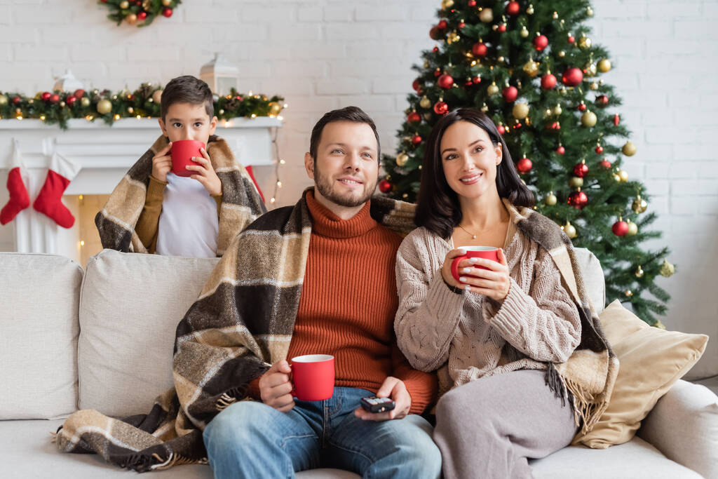 uśmiechnięta rodzina z ciepłymi kocami trzymając filiżanki kakao w salonie z wystrojem świątecznym - Zdjęcie, obraz