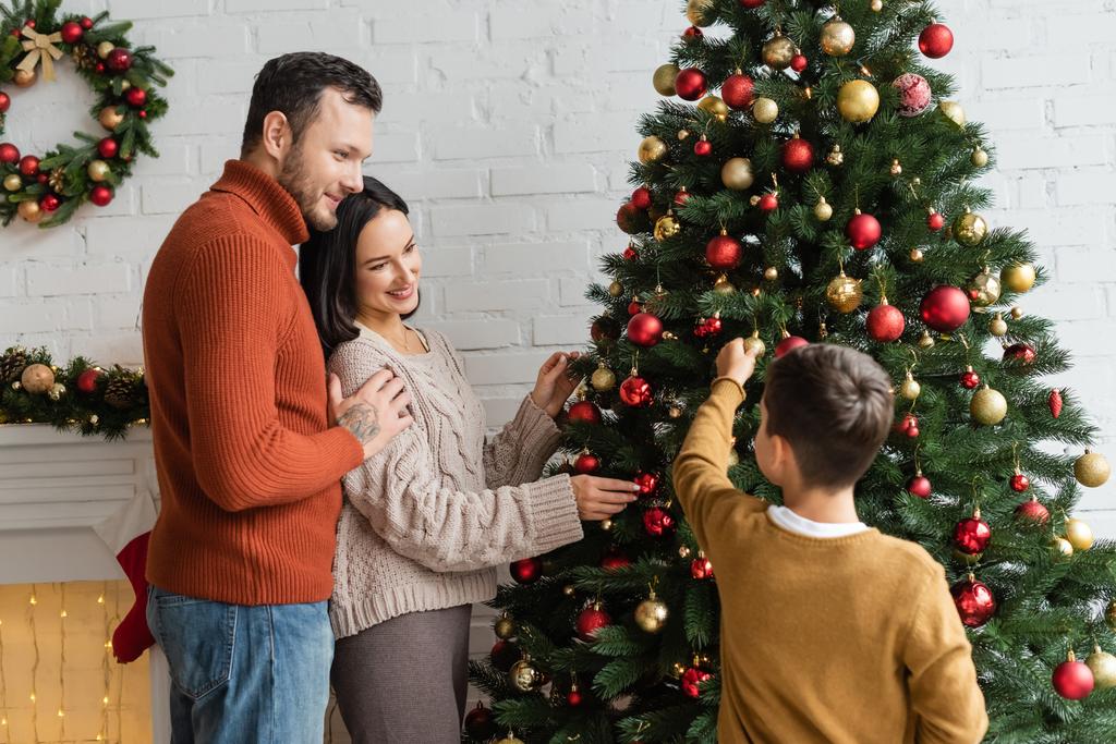 uomo sorridente abbraccio allegro moglie decorare abete rosso verde con baubles Natale vicino al figlio - Foto, immagini