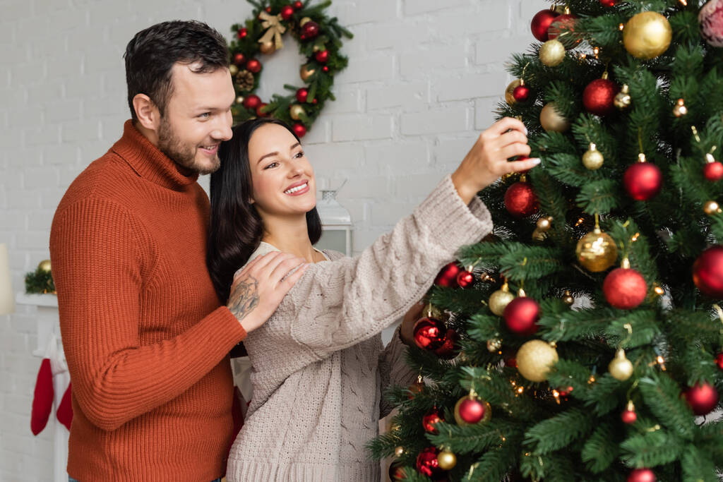 smiling man embracing happy and pretty wife decorating green spruce with christmas baubles - Photo, Image