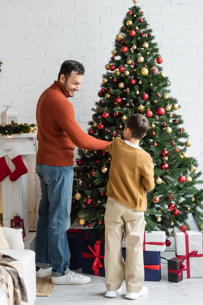 wesoły ojciec z synem dekorowanie zielonej sosny z bożonarodzeniowych kulek w pobliżu pudełek prezentów - Zdjęcie, obraz