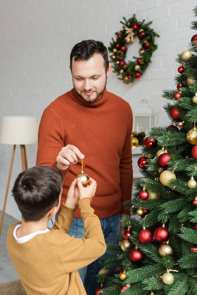 smiling dad giving christmas ball to son decorating green spruce at home - Photo, Image