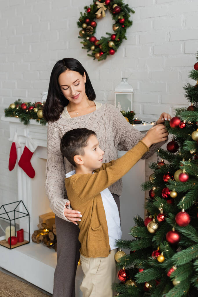 усміхнена жінка допомагає синові прикрашати зелену сосну з різдвяними камінчиками вдома
 - Фото, зображення