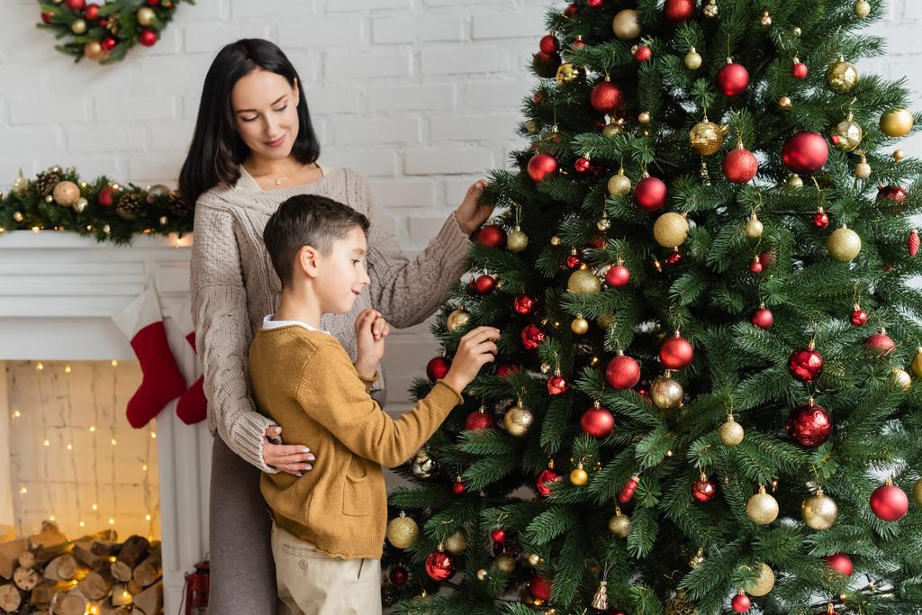šťastná žena objímající syna zdobení vánoční strom v blízkosti krbu v obývacím pokoji - Fotografie, Obrázek