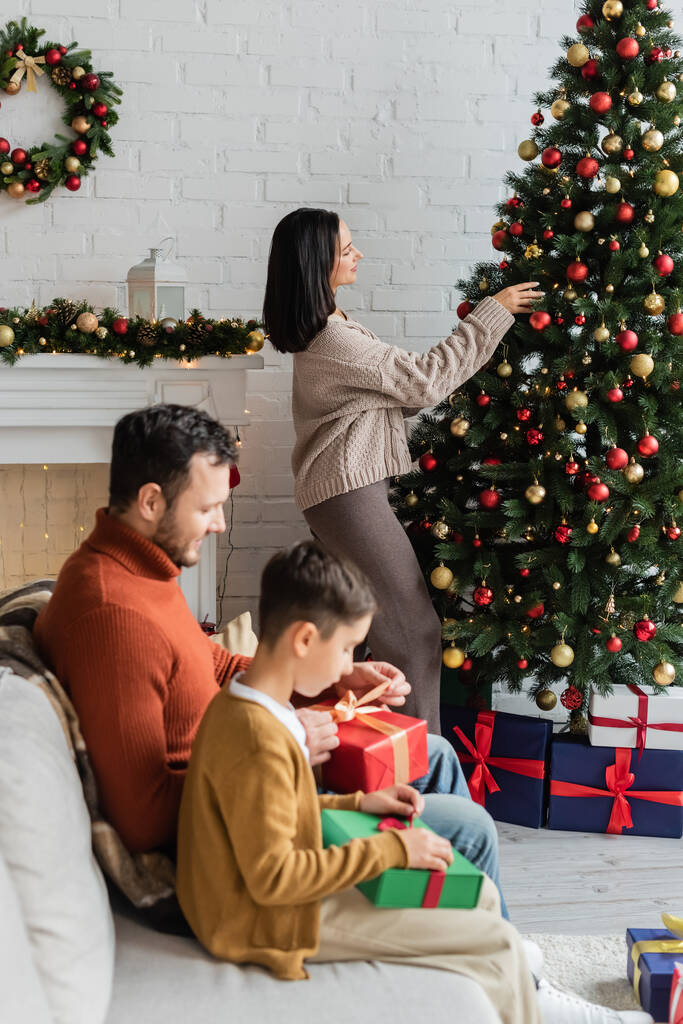 oldal kilátás nő díszítő karácsonyfa közelében férj és fia csomagolás ajándékok kanapén - Fotó, kép