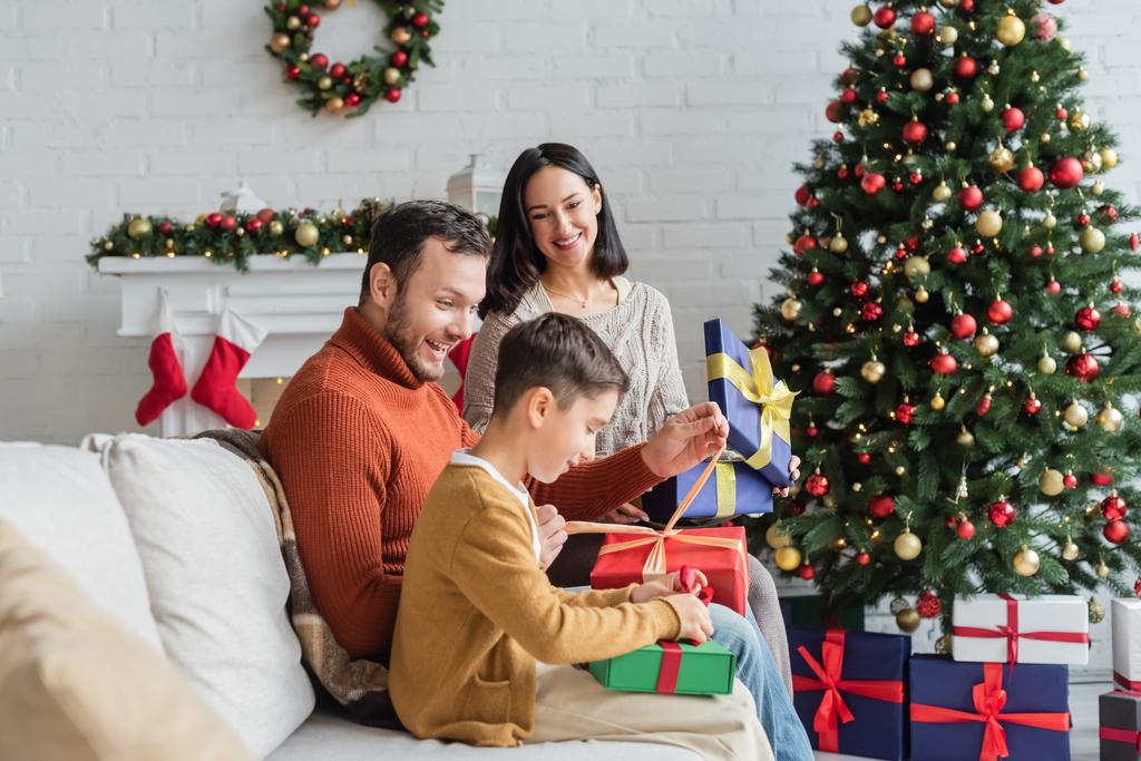 vidám család csomagolási ajándék dobozok kanapén közel karácsonyfa és díszített kandalló - Fotó, kép
