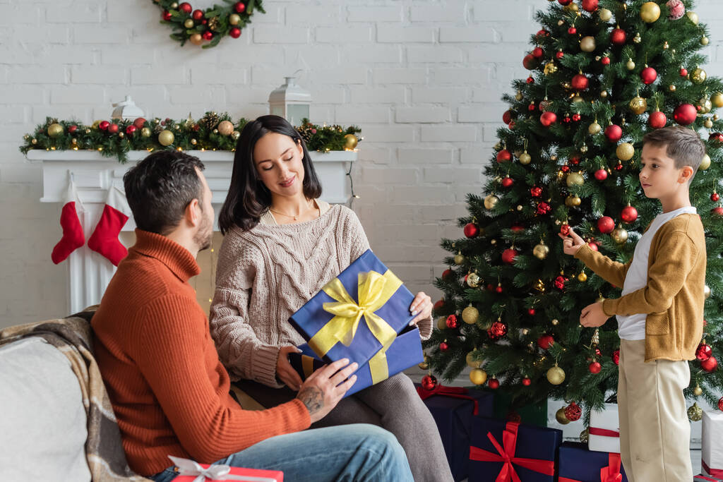 sonriente esposa abriendo caja de regalo cerca de marido e hijo decorando árbol de Navidad - Foto, imagen