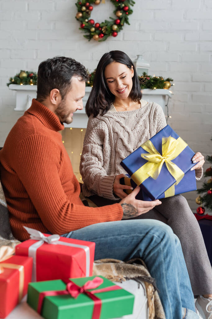 Lächelnde Frau mit Weihnachtsgeschenk, während sie auf der Couch neben Ehemann und Geschenkschachteln sitzt - Foto, Bild
