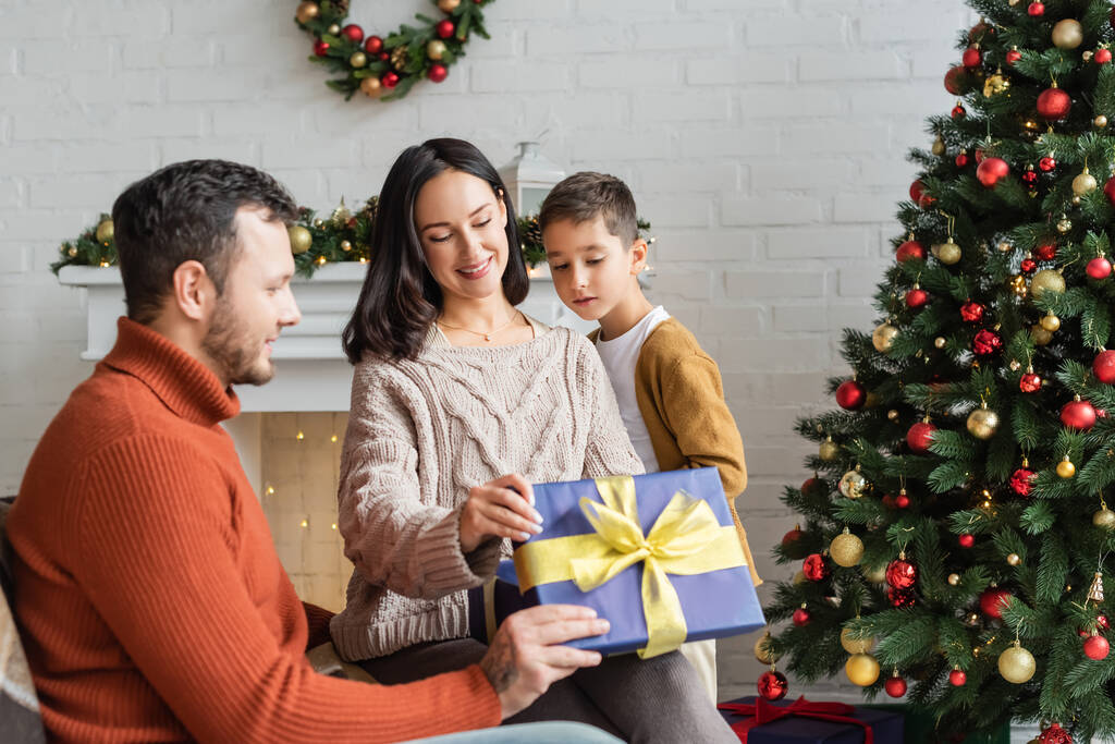 happy woman holding gif box near husband and son in living room with decorated christmas tree - Photo, Image