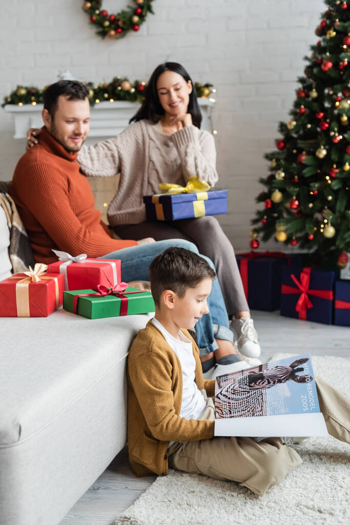 Lächelnder Junge liest Magazin auf dem Boden neben verschwommenen Eltern und Weihnachtsgeschenken auf der Couch im Wohnzimmer - Foto, Bild