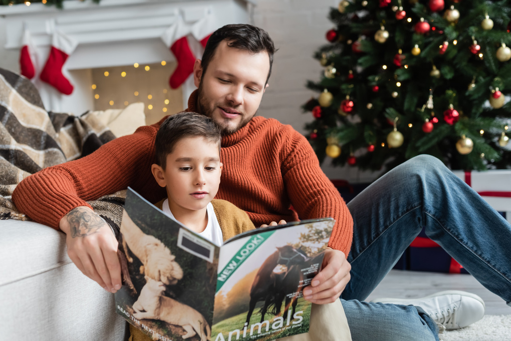 menino leitura revista com sorrindo pai perto borrada árvore de Natal - Foto, Imagem